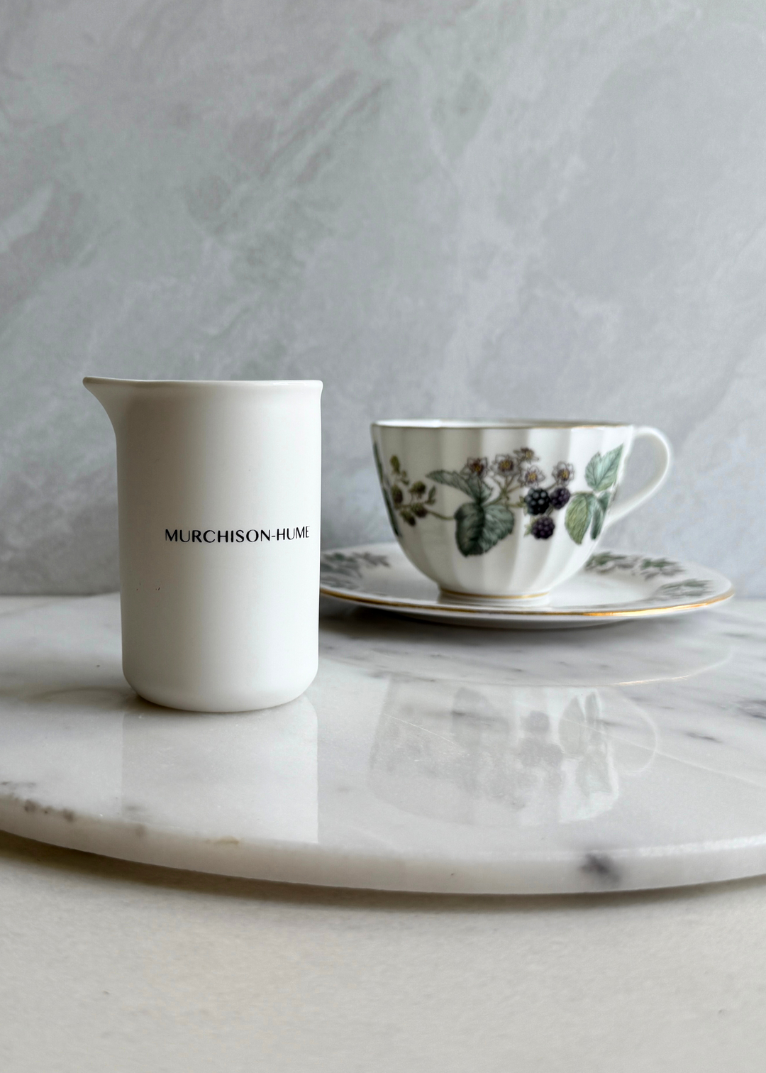 White pitcher with 'Murchison Hume' branding next to a floral teacup on a marble surface.